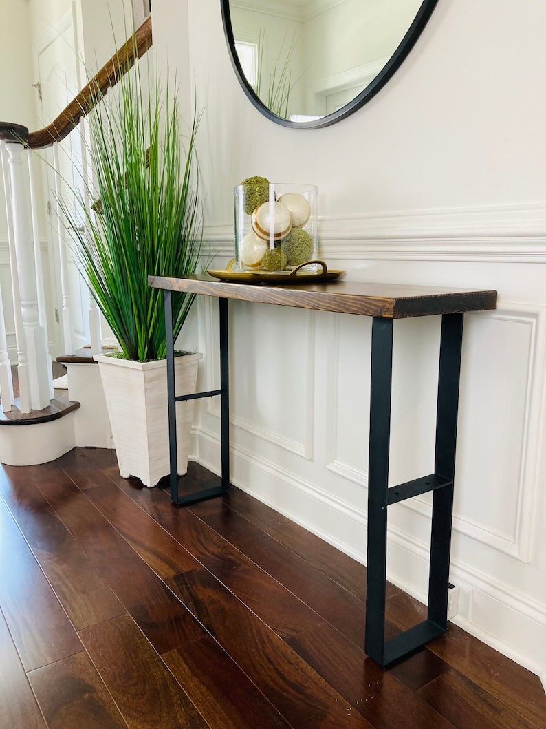 May include: A wooden console table with black metal legs. The table has a dark wood finish and is decorated with a vase of green plants and a glass jar filled with decorative balls.