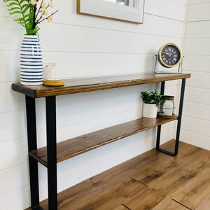 Puede incluir: Una mesa consola de madera con un marco de metal negro y dos estantes. La mesa tiene un acabado r&uacute;stico y desgastado y est&aacute; decorada con un jarr&oacute;n de flores, un reloj y una planta en maceta.