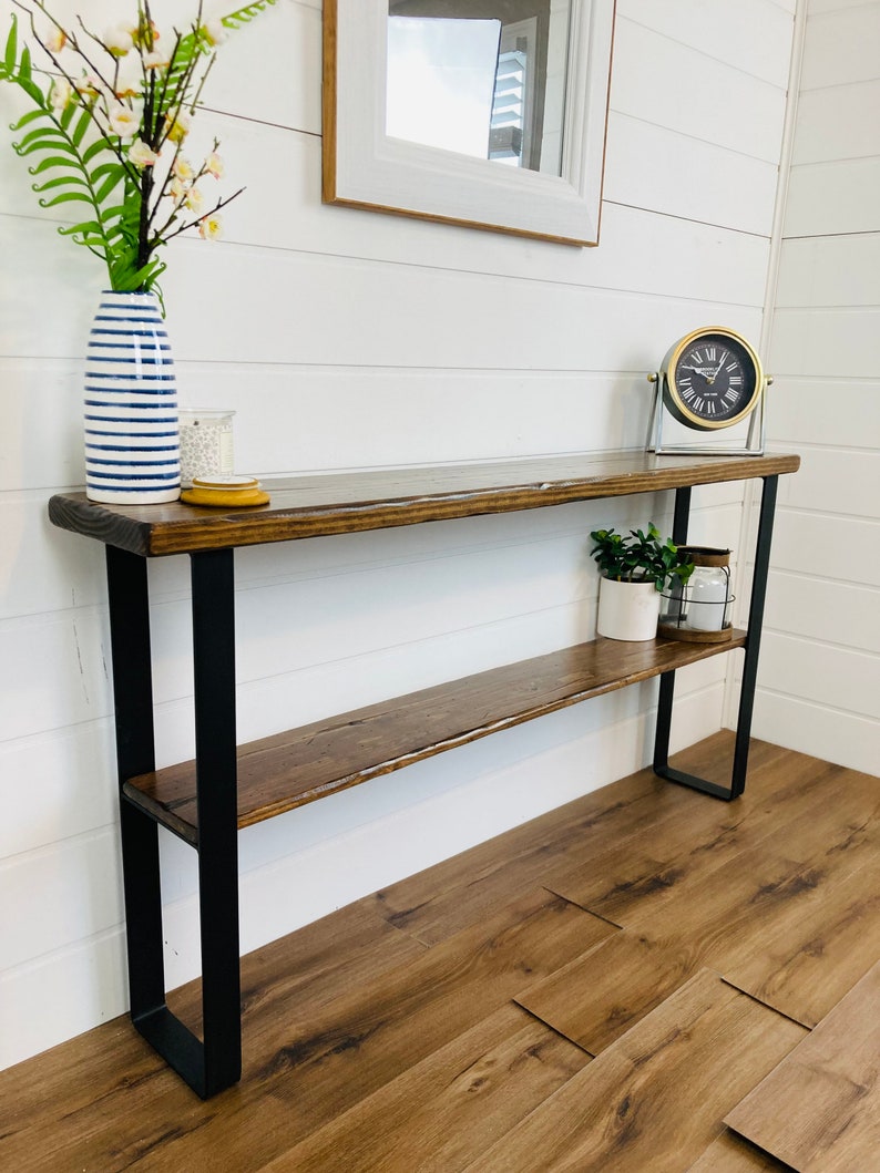 Puede incluir: Una mesa consola de madera con un marco de metal negro y dos estantes. La mesa tiene un acabado r&uacute;stico y desgastado y est&aacute; decorada con un jarr&oacute;n de flores, un reloj y una planta en maceta.