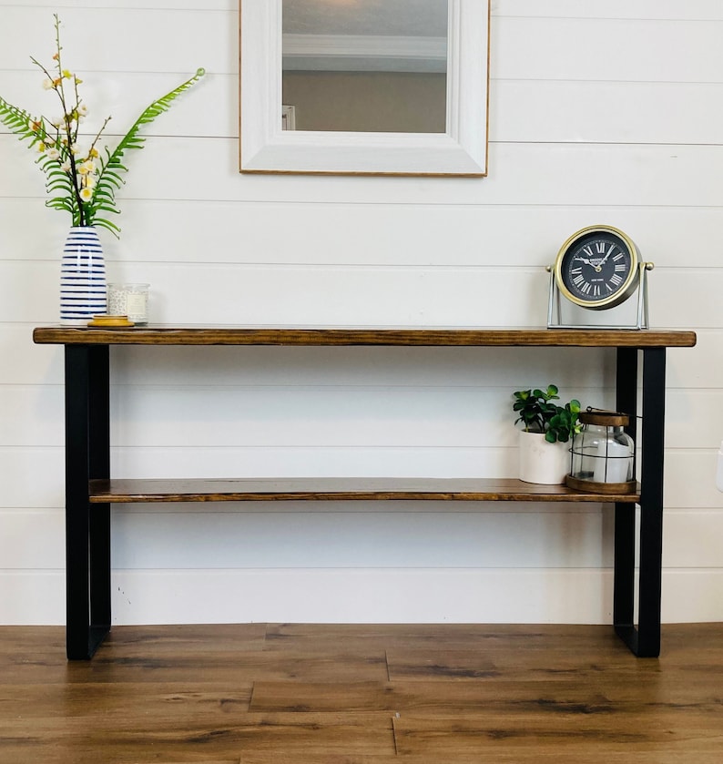 Puede incluir: Una mesa consola de madera marr&oacute;n oscuro con patas de metal negro y un estante inferior. La mesa tiene un reloj dorado en el estante superior y una planta en maceta en el estante inferior.