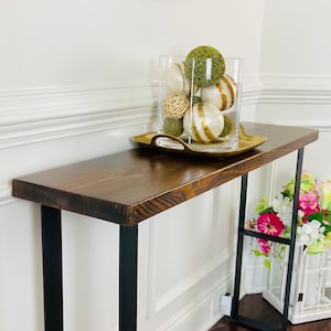 May include: A dark brown wooden console table with black metal legs. The table has a distressed finish and is decorated with a glass vase filled with green and white decorative balls. The vase is sitting on a gold tray.
