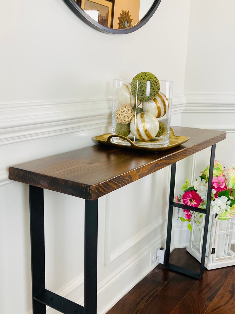May include: A dark brown wooden console table with black metal legs. The table has a distressed finish and is decorated with a glass vase filled with green and white decorative balls. The vase is sitting on a gold tray.