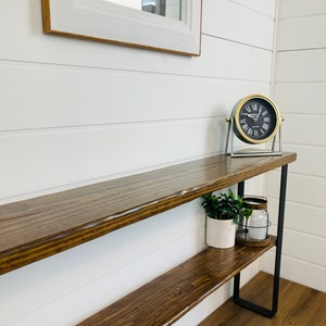 Puede incluir: Una mesa consola de madera con dos estantes y patas de metal negro. La mesa est&aacute; decorada con una peque&ntilde;a planta en maceta, un portavelas y un reloj redondo con n&uacute;meros romanos.