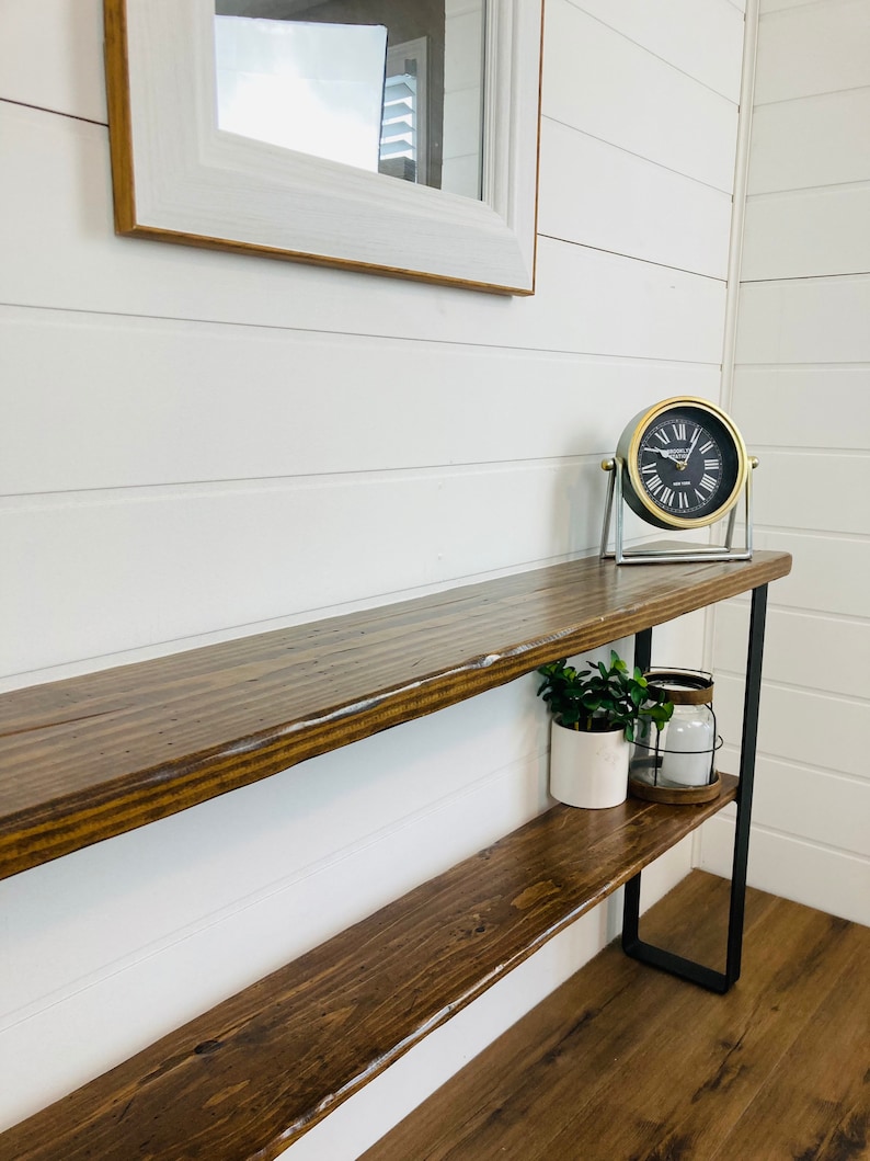Puede incluir: Una mesa consola de madera con dos estantes y patas de metal negro. La mesa est&aacute; decorada con una peque&ntilde;a planta en maceta, un portavelas y un reloj redondo con n&uacute;meros romanos.