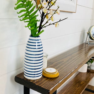 Puede incluir: Una mesa consola de madera con patas de metal negro. La mesa tiene dos estantes y est&aacute; decorada con un jarr&oacute;n de rayas azules y blancas lleno de flores blancas y una vela amarilla.