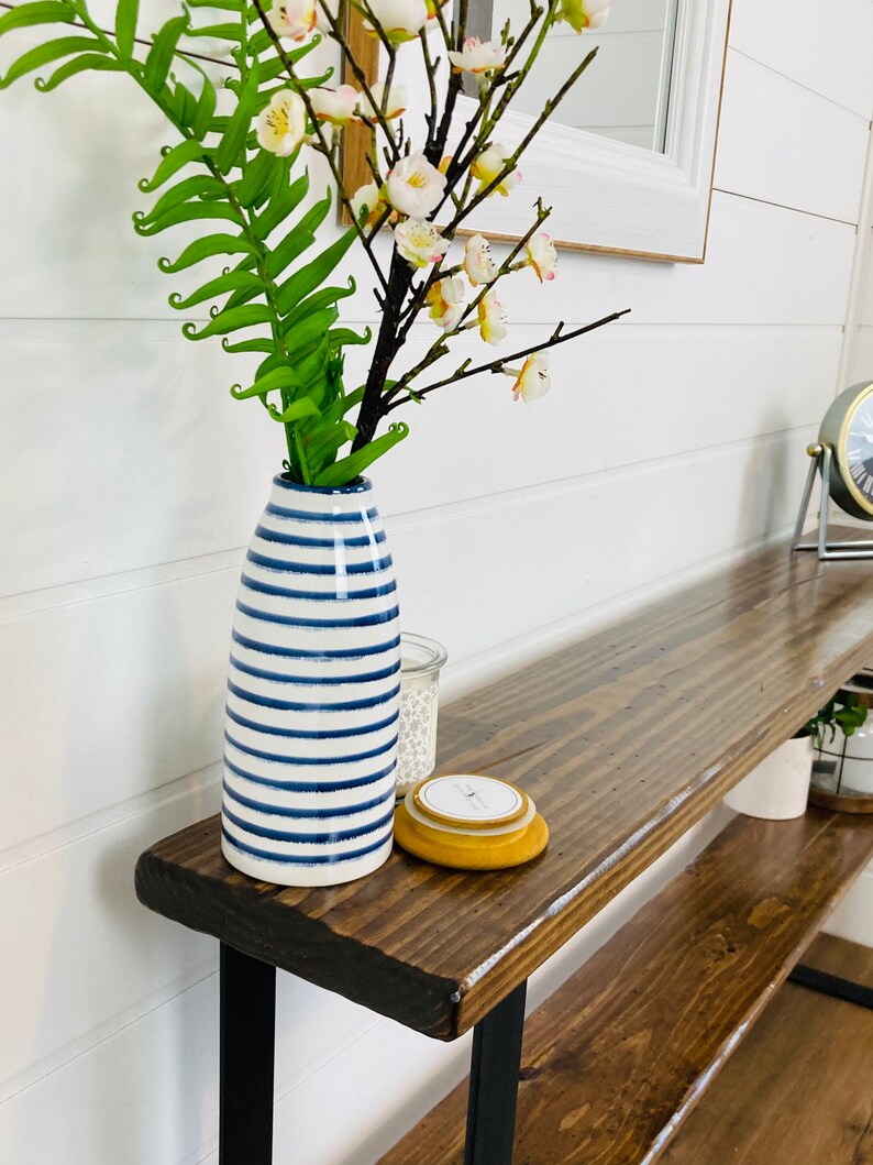 Puede incluir: Una mesa consola de madera con patas de metal negro. La mesa tiene dos estantes y est&aacute; decorada con un jarr&oacute;n de rayas azules y blancas lleno de flores blancas y una vela amarilla.