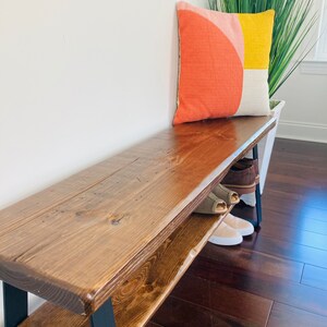 May include: A wooden bench with a black metal frame and a shelf underneath. The bench has a dark brown stain and is topped with a colorful geometric pillow.