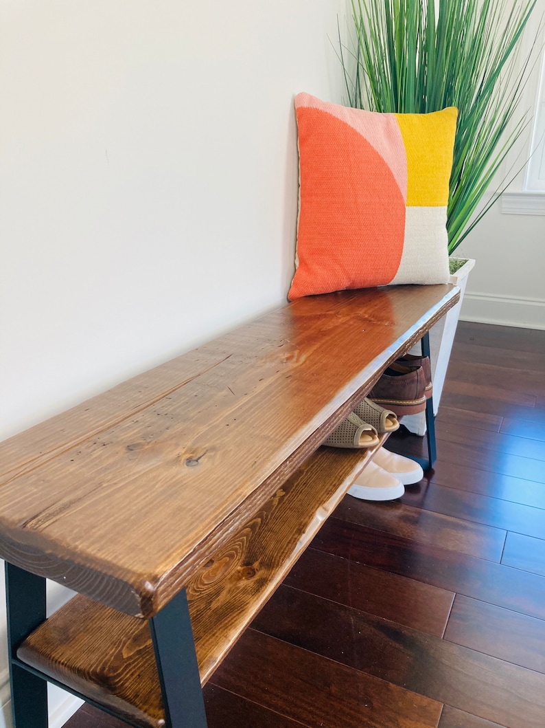 May include: A wooden bench with a black metal frame and a shelf underneath. The bench has a dark brown stain and is topped with a colorful geometric pillow.
