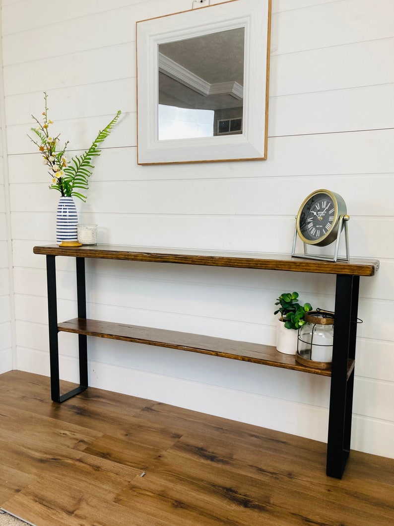 Puede incluir: Una mesa consola de madera marr&oacute;n oscuro con patas de metal negro y un estante. La mesa tiene un jarr&oacute;n blanco con flores y un reloj en la parte superior.