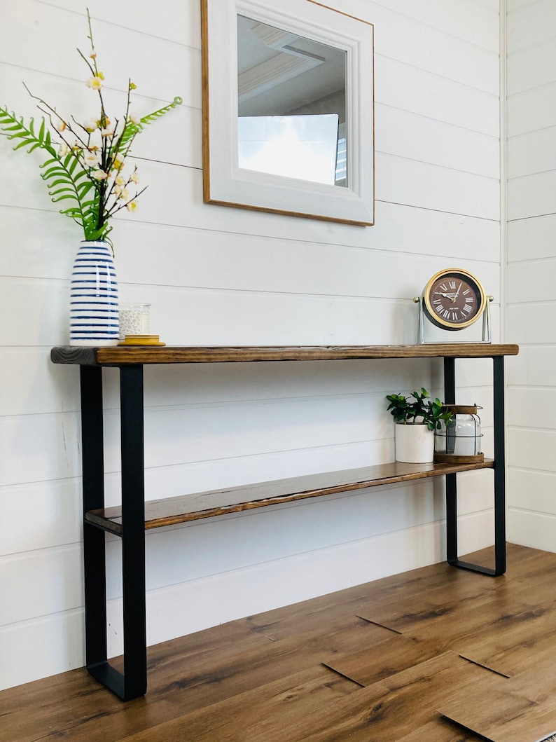 Puede incluir: Una mesa consola de madera con patas de metal negro y dos estantes. La mesa tiene un acabado de madera envejecida y est&aacute; decorada con un jarr&oacute;n de flores, un reloj y una planta en maceta.