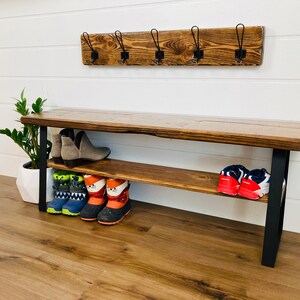 May include: A wooden bench with a shelf for shoes, featuring black metal legs and a rustic brown finish. The bench has a coat rack above it with five hooks.