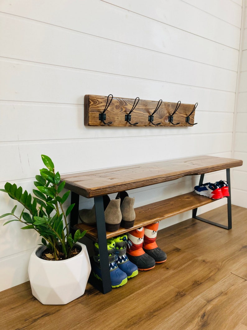 May include: A wooden bench with a metal frame and two shelves for storing shoes. The bench has a rustic brown finish and the metal frame is black. There are hooks on the wall above the bench.