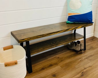 May include: A wooden bench with a black metal frame and a lower shelf. The bench has a dark wood stain and is sitting on a light wood floor. A white and blue pillow is on the bench.