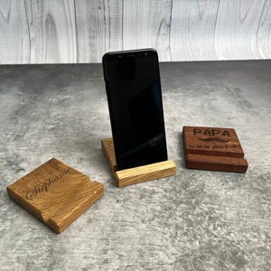 Peut inclure: Trois supports de téléphone en bois, l'un avec un smartphone noir, l'autre avec le texte "Stephanie" et le dernier avec le texte "PAPA Tu es le plus fort!"