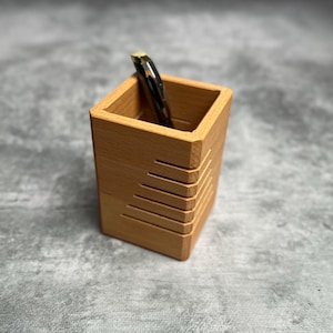 Peut inclure: Un petit porte-stylo en bois brun clair avec un motif décoratif sur un côté. Un stylo noir et doré se trouve à l'intérieur du support. Le porte-stylo est carré et repose sur une surface grise. C'est un accessoire de bureau.