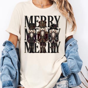 May include: A cream-colored short-sleeve t-shirt featuring a graphic print of three nutcracker figures in shades of red, black, and gold. The text MERRY MERRY MERRY is printed in a large black serif font behind the nutcracker illustrations.