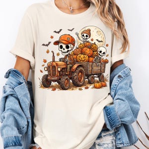 May include: A cream-colored t-shirt featuring a graphic of skeletons driving a tractor pulling a wagon filled with pumpkins. The scene includes a full moon, bats, and falling autumn leaves. The skeleton driver wears an orange cap and plaid shirt.