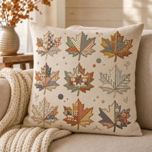 May include: A square throw pillow with a cream-colored fabric cover featuring a grid of nine embroidered maple leaves. Each leaf displays a unique patchwork pattern in autumn colors including orange, brown, blue, and yellow, accented by small buttons.