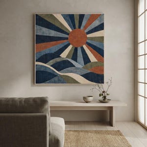 May include: A square wall art piece featuring a textured sunburst design with a central rust-colored circle, radiating beams in shades of navy blue, olive green, cream, and rust, set above stylized rolling hills in deep blue and muted green tones.