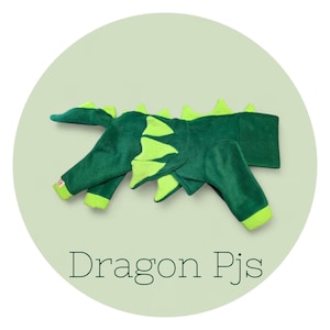 Puede incluir: Pijama de dragón verde con detalles en verde lima. El pijama tiene un diseño de escamas de dragón en la espalda y una cola. El texto "Dragon Pjs" está impreso en la parte inferior de la imagen.