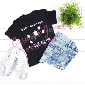 May include: Black t-shirt with white text that reads "Best Dentist in the Molar System". The text is surrounded by a graphic of teeth in a circular pattern. The t-shirt is laid out on a white wooden surface with a pair of white sneakers, a pair of blue denim shorts, and a potted green plant.