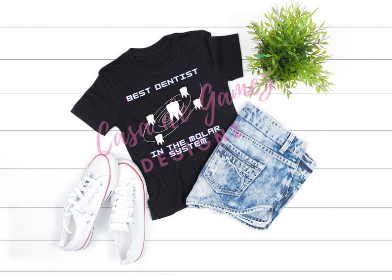 May include: Black t-shirt with white text that reads "Best Dentist in the Molar System". The text is surrounded by a graphic of teeth in a circular pattern. The t-shirt is laid out on a white wooden surface with a pair of white sneakers, a pair of blue denim shorts, and a potted green plant.