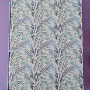 Può includere: Un diario con un motivo blu chiaro e verde su sfondo bianco. Il diario ha una rilegatura semplice con filo blu.