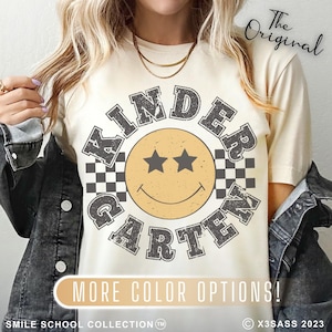 May include: Cream-colored t-shirt with a retro "Kindergarten" design featuring a smiley face with star eyes and a checkered border. The text "MORE COLOR OPTIONS!" is below the design. The words "The Original" are at the top.