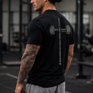 A man wearing a black t-shirt with a graphic design on the back, standing in a gym.