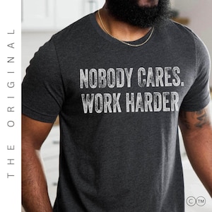 Puede incluir: Camiseta gris oscuro con el texto blanco "NOBODY CARES. WORK HARDER". El texto tiene un efecto desgastado. La camiseta tiene cuello redondo y mangas cortas. Se ve una cadena dorada.
