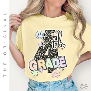 以下が含まれることがあります： 「4th GRADE」と書かれたカラフルなグラフィックが特徴の、明るい黄色のTシャツ。デザインには星、スマイリーフェイス、「th」の文字が含まれています。このTシャツはカジュアルなスタイルです。