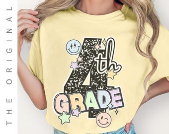 May include: A light yellow t-shirt with a colorful graphic that says "4th GRADE". The design includes stars, smiley faces, and the word "th". The shirt is a casual style.