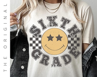 May include: Cream-coloured Sixth Grade t-shirt featuring a retro smiley face design. The smiley face is yellow with two star eyes and a smiling mouth. The words "SIXTH GRADE" are in a distressed, grey font, with checkered flag accents.