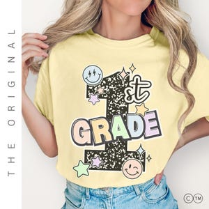 以下が含まれることがあります： パステルカラーの「1st GRADE」の文字がプリントされた、薄い黄色のTシャツ。数字の1は黒で、白い縁取りと斑点模様。スマイリーフェイスと星がシャツを飾っています。