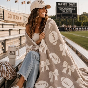 May include: A beige and white plush throw blanket featuring a repeating pattern of footballs and bows. The blanket is draped over a person wearing a beige baseball cap with the text SUNDAY FUNDAY, a white top, and blue jeans, next to a tumbler.