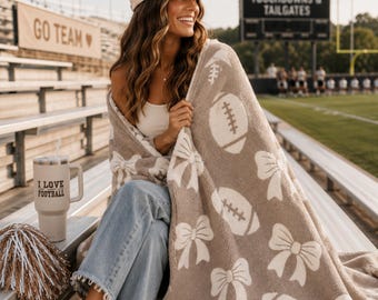 Puede incluir: Una manta de felpa beige y blanca con un patrón repetitivo de balones de fútbol americano y lazos. La manta está colocada sobre una persona que lleva una gorra beige con el texto SUNDAY FUNDAY, una camiseta blanca y pantalones vaqueros azules.