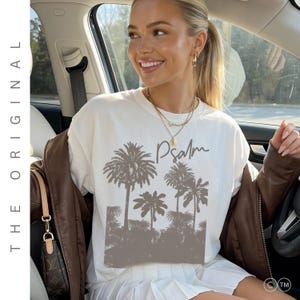 May include: A white short-sleeved t-shirt featuring a dark brown graphic print of palm trees and the word Psalm written in a cursive script. The shirt is worn with a brown jacket and a white pleated skirt, accessorised with a gold necklace.