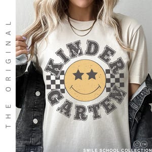 May include: Cream-colored t-shirt with a retro "Kindergarten" design. The design features a yellow smiley face with star eyes, surrounded by the words "Kindergarten" in a distressed gray font, with checkered accents.