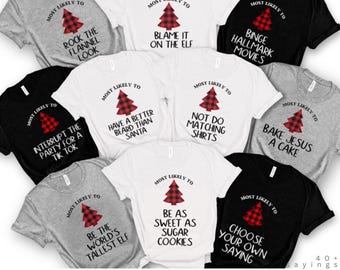 May include: A collection of t-shirts in various colors, each featuring a red and black plaid Christmas tree graphic. The shirts have phrases like "Most Likely To Rock The Flannel Look" and "Choose Your Own Saying."