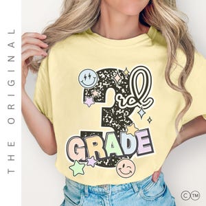 May include: A light yellow t-shirt with a colorful graphic that says "3rd Grade". The graphic includes a smiley face, stars, and the word "GRADE" in pastel colors. The text "THE ORIGINAL" is printed vertically on the left side.