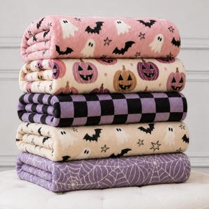 May include: A stack of five folded fleece blankets. The top blanket is pink with white ghosts, black bats, and stars. The second is cream with purple and orange jack-o'-lanterns. The third is a purple and black checkerboard. The fourth is cream with ghosts, bats, and stars. The bottom is purple with a white spiderweb pattern.