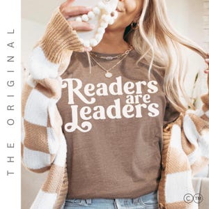 Könnte beinhalten: Ein braunes T-Shirt mit dem Text "Readers are Leaders" in weißer Schrift. Die Person trägt eine karierte Strickjacke in Beige und Weiß sowie goldene Halsketten. Das Bild hat den Text "THE ORIGINAL" auf der linken Seite.