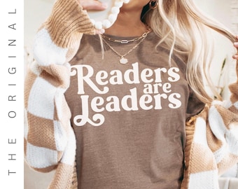 May include: A brown t-shirt with the text "Readers are Leaders" in white script. The person is wearing a tan and white checkered cardigan and gold necklaces. The image has the text "THE ORIGINAL" on the left side.