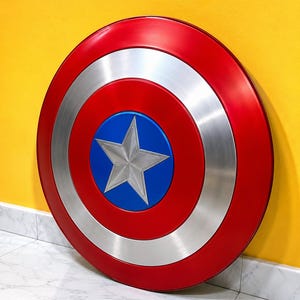 Puede incluir: Un escudo redondo rojo, plateado y azul, con una estrella plateada en el centro sobre un disco azul. El escudo tiene un anillo exterior rojo y una banda plateada. Un accesorio ic&oacute;nico de superh&eacute;roe.