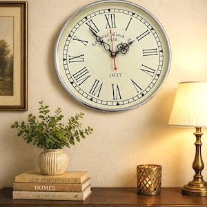 Puede incluir: Un reloj de pared redondo con marco plateado y esfera crema. Tiene números romanos negros, manecillas negras y un segundero rojo. El texto "Colonial Clock estd. 1937" está impreso en la esfera.