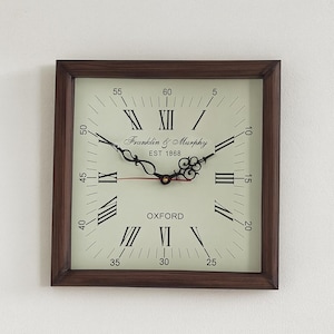 May include: Square wall clock with a dark brown wooden frame. The clock face is off-white with black Roman numerals and hands. The clock reads "Franklin & Murphy EST 1968 OXFORD".