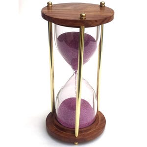 Könnte beinhalten: Eine Sanduhr mit einem Holzdeckel und -sockel, verbunden durch messingfarbene Metallstäbe. Die Glaskammer enthält violetten Sand. Die Sanduhr ist ein dekorativer Gegenstand.