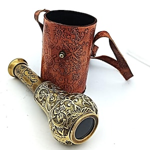 May include: A brass antique spyglass with floral engravings, next to a brown leather case with nautical-themed embossing. The spyglass has a black lens and a gold finish, evoking a vintage aesthetic.