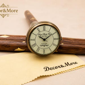 May include: A vintage-style clock with a cream-colored face and black Roman numerals. The clock is set in a brass-colored casing and attached to a wooden handle. The text "Royal Clocks Co. Est. 1842 Scotland" is visible on the clock face. A yellow cloth and the text "Decor&More" are in the foreground.