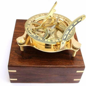 May include: A vintage brass sundial compass with Roman numerals and a degree scale, mounted on a dark wooden box. The compass features a polished gold finish and is designed for navigation and timekeeping. The box has brass accents.